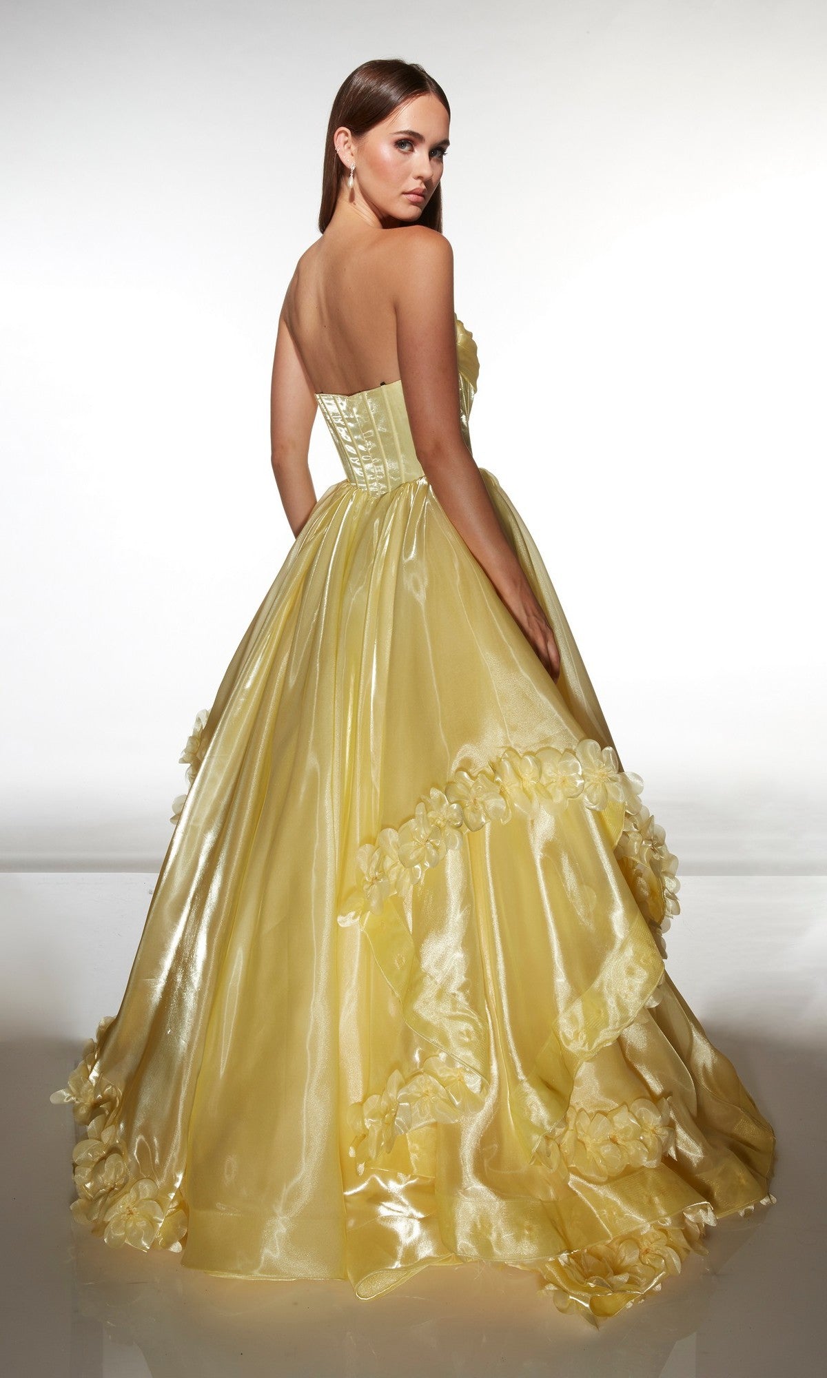 A woman wearing the Alyce 62014 Long Prom Dress, a strapless ballgown in liquid organza with 3D floral appliques, stands facing away and glances over her shoulder.